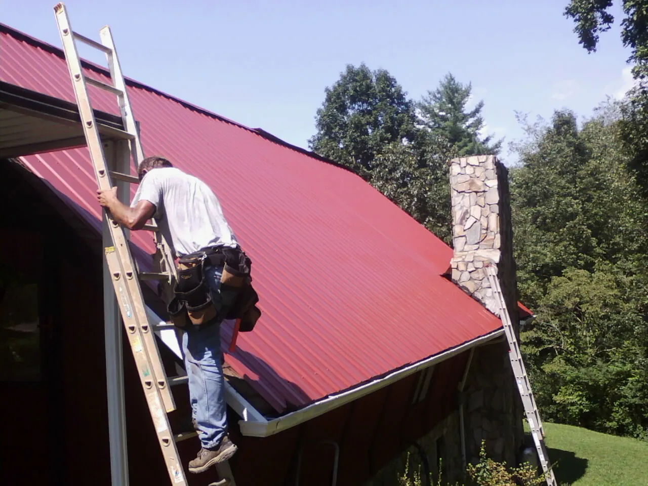 Roofer on a ladder beside a red metal roof and stone chimney — Soffit & Fascia Repair by Roofing Brothers in Paola